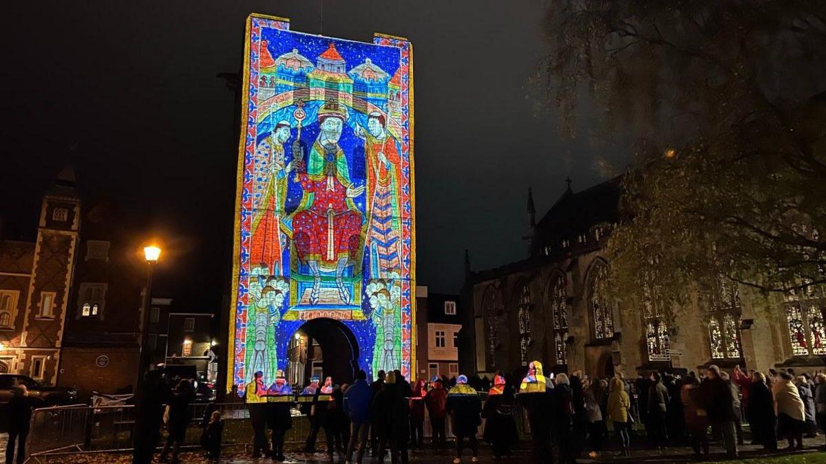 A crowd gathered around a colourful projected image of St Edmund at St Edmundsbury Cathedral. It resembles a gigantic stained-glass window.

