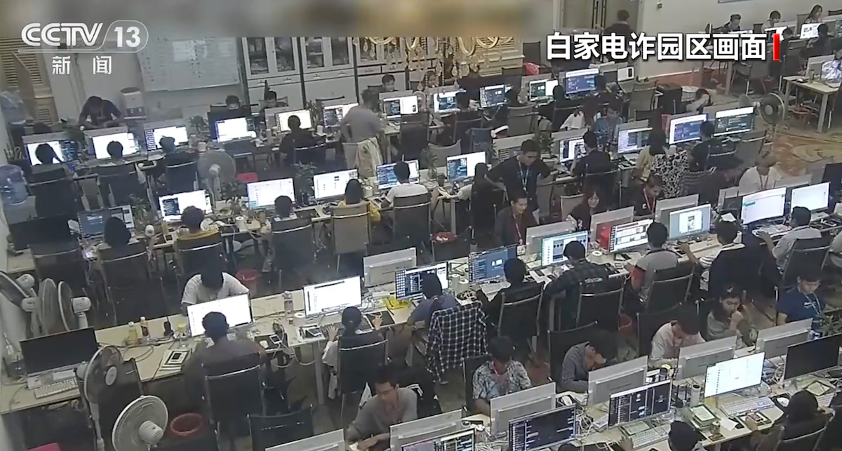 Screenshot from a CCTV documentary showing security camera footage of a scam centre, with workers sitting at rows of chairs, each looking at their computer screens.