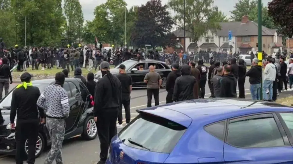 Crowds gather on a street near a McDonald's close to the Heartlands Hospital, with three cars in the shot
