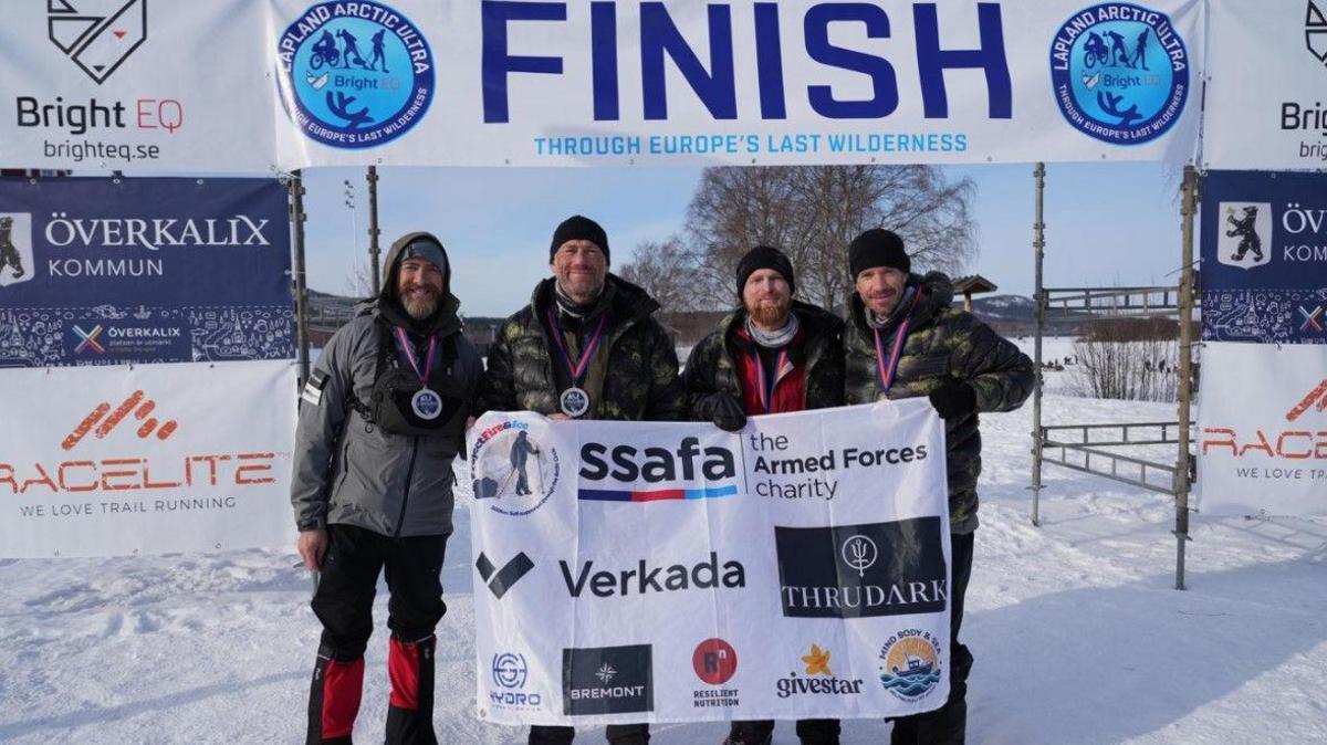 Aldo Kane, Jason Fox, Karl Hinett and author Brian Wood (left to right) in snow at the finish of the ultra marathon. The word finish is in large letters above them. They are holding up a white sheet including the name SSAFA, the armed forces charity.
 