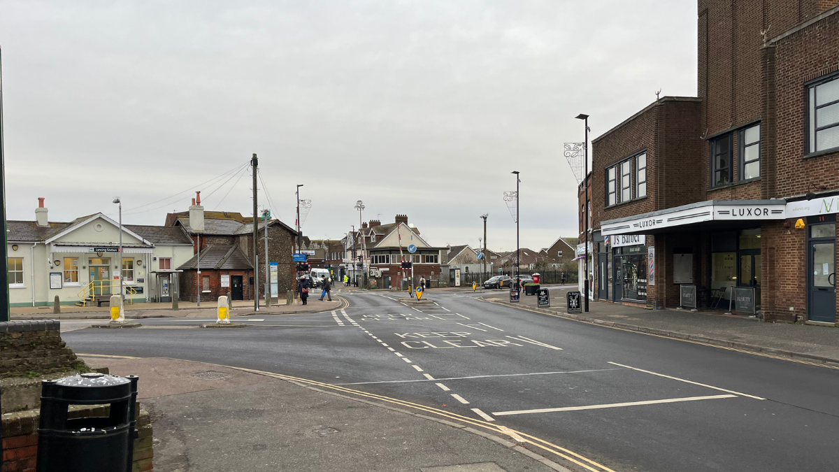 South Street looking towards Lancing railway station, there's a junction veering to the left. A tall brown building on the right, and the crossing can be seen in the distance. The road and pavements are almost completely empty