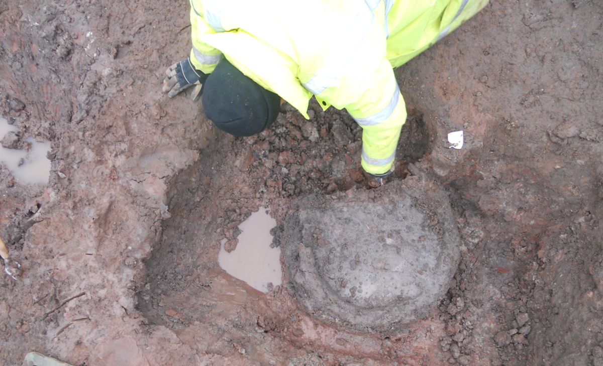 A man in hi-vis clothing pulls a round object from a hole in mud