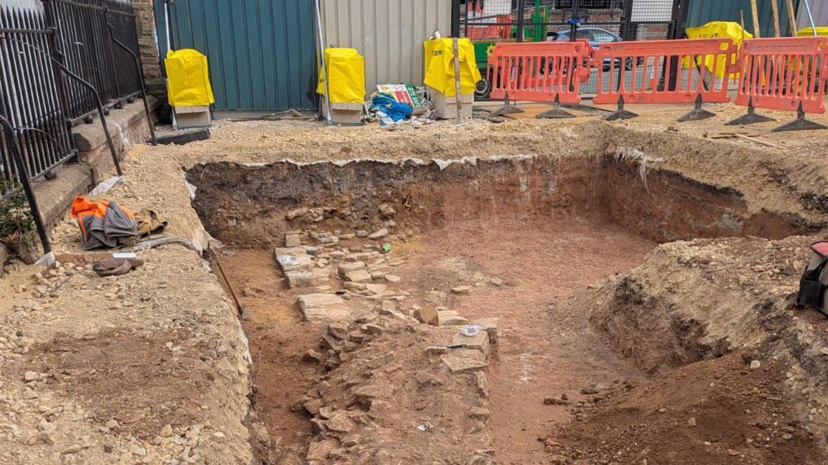 A pit carved out of earth with bricks inside and equipment and orange plastic fencing around the egde.