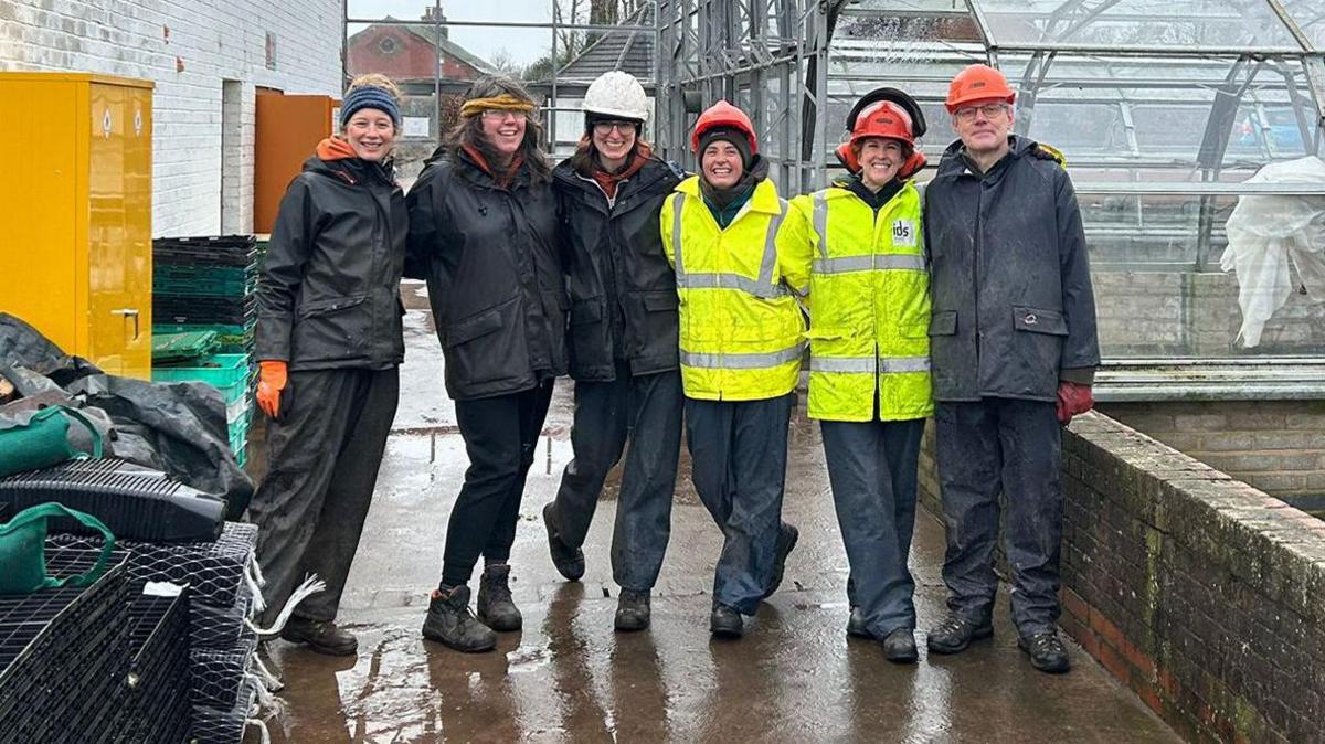 Five people, some wearing hard hats and hi-vis jackets stand in front of a greenhouse. It is raining and they are all wearing waterproof clothing 