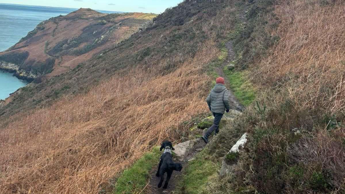 Solly is walking with his back to the camera along a trail path on the north coast of Jersey. Behind him, a black dog is following him. Solly is wearing blue trousers, a dull green thick coat and a dull pink woolen hat.