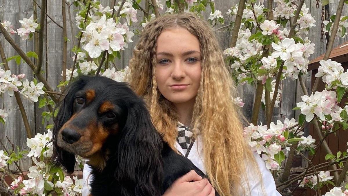 Lily-May pictured looking into the camera and smiling slightly. She's wearing a white jumper and is holding her dog Sadie, a black and tan cocker spaniel. Behind her is a tree covered in white flowers, and beyond that, a panelled wood fence.
