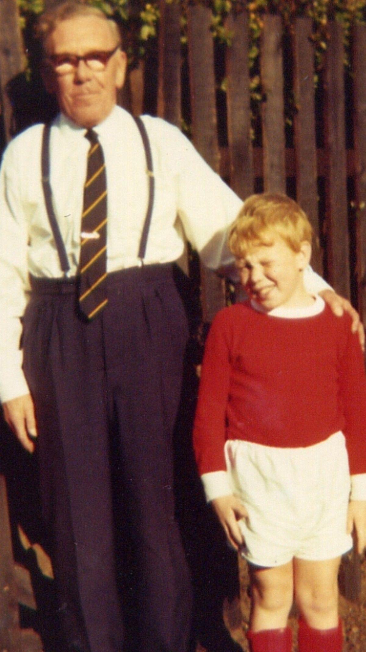 An older man dressed in a suit and tie stands with a younger boy dressed in a red and white football kit