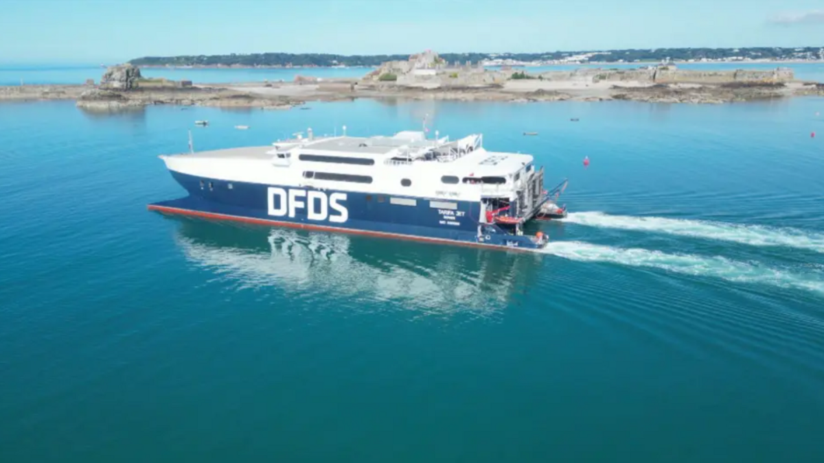 A wide shot of a DFDS ferry out on the water. It is painted white on top and blue below. The water is calm and it is a sunny day.