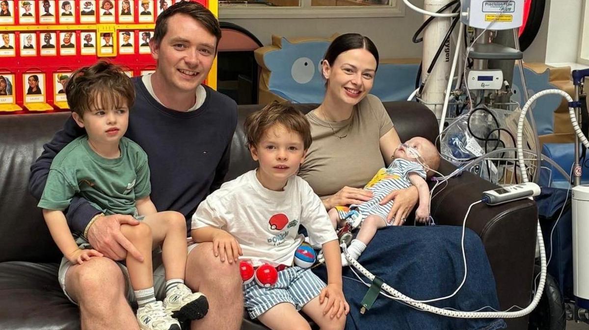 Macs Green and family. His family are sat on a sofa. Macs is on his mother's lap. He is connected to hospital machinery