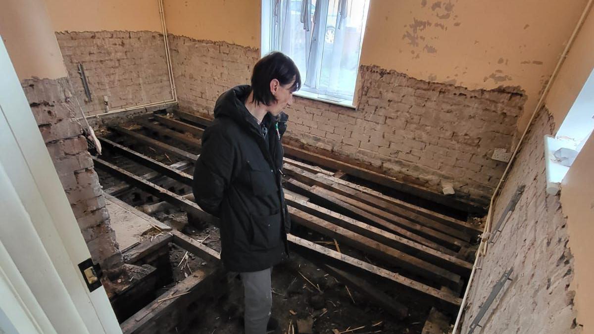 Carlie looking through the floorboards of her flooded living room, with exposed brick on the walls