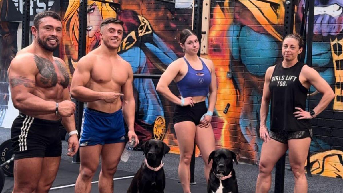 Two bare-chested men, one in black shorts, the other in blue shorts and two women in sports gear standing in a row with two black dogs in sitting on the ground in front of them