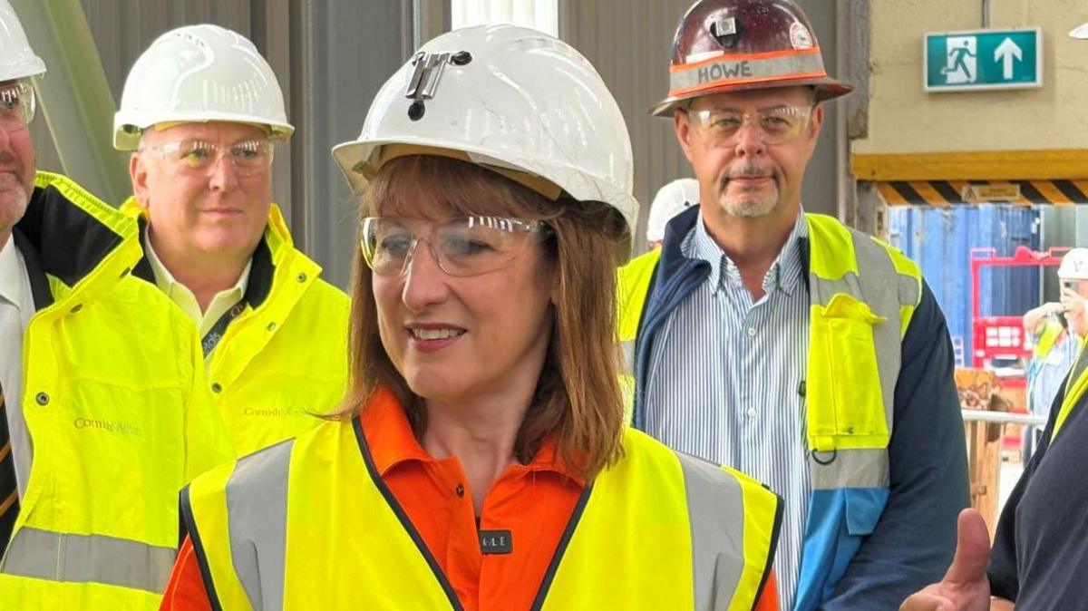 Chancellor Rachel Reeves is wearing an orange top and yellow high vis jacket and a white hard hat. Several men who are wearing similar protective clothing are standing behind her.
