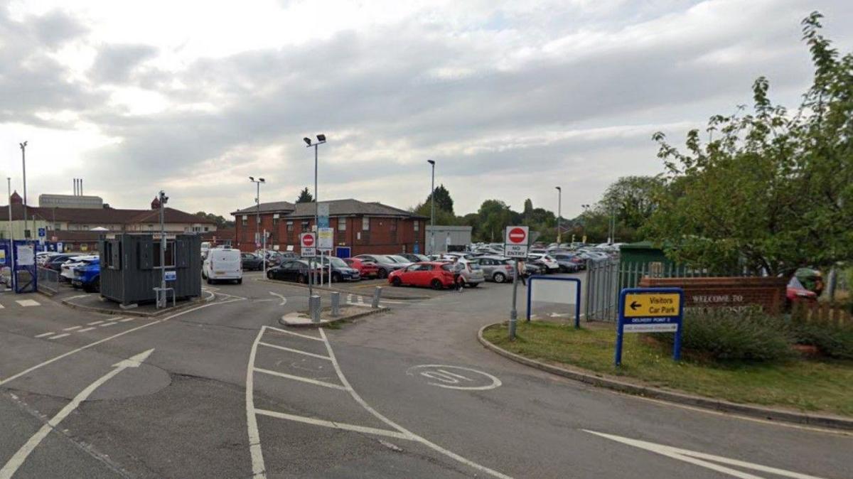 Hospital site with buildings in the background. One lane leads to a car park and another goes in the opposite direction for vehicles leaving the site. A Warwick Hospital sign is partially obscured by foliage on the right of the photo.