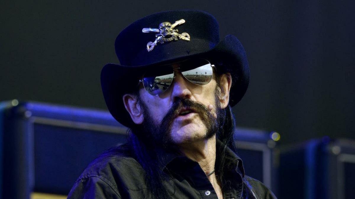 A head and shoulders shot of the singer in a black shirt, dark glasses and black hat with a silver skull and cross bones on a stage. He has a dark handlebar moustache.