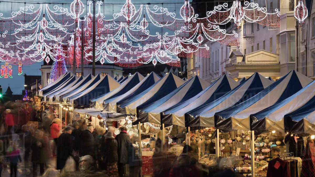 The Christmas market in Stratford-upon-Avon two years ago, with people wearing coats and hats and carrying bags walking past stalls, which have blue canopies over them and there are Christmas lights overhead.