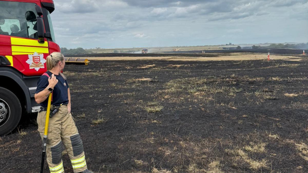 A firefighter stands in a burnt field holding a shovel, with a fire engine nearby. The sky is partly cloudy, and the landscape in the background shows more burnt areas.