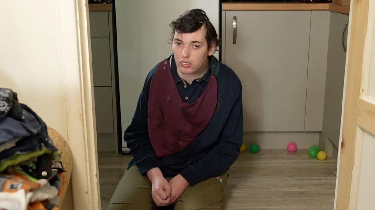 Harry kneels on the floor within a door frame. He looks away from the camera. He wears a blue jumper with green trousers and a red bib.