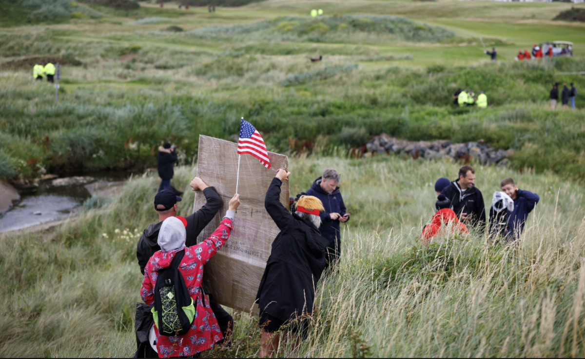 Three of protesters holding an American flag and a placard in the grounds of Turnberry with their backs to the camera. They are standing in a tuft of long grass with a golf course stretching in front of them. The resort's buildings can be seen in the background.