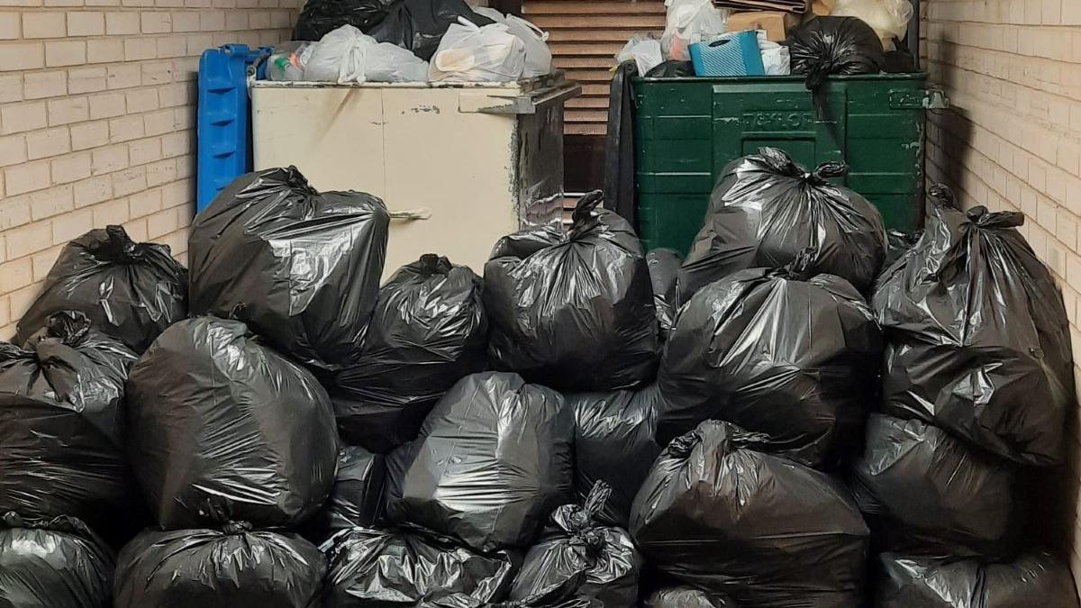 Piles of black bin bags next to two large bins which are overflowing with rubbish.