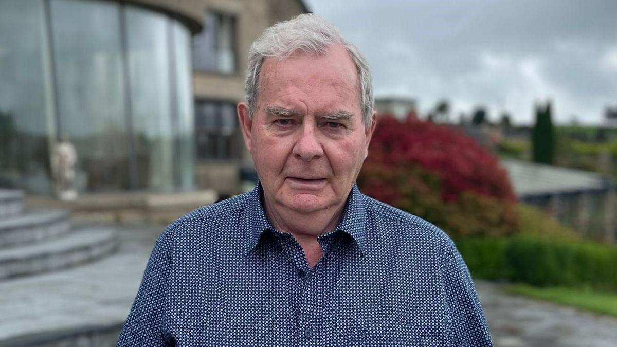 Seán Quinn - a man with short grey hair looks at the camera with his mouth open. He is wearing a blue patterned shirt with the top button undone. He is standing outside of a home with large windows, but the background is blurred.