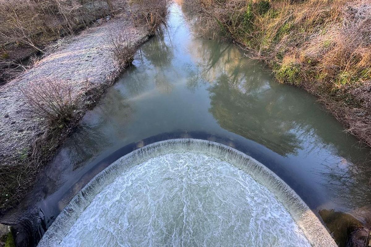 An aerial view of a weir in Downton Gorge. Water flows over the horseshoe shaped weir while upstream the river banks banks are covered in frost