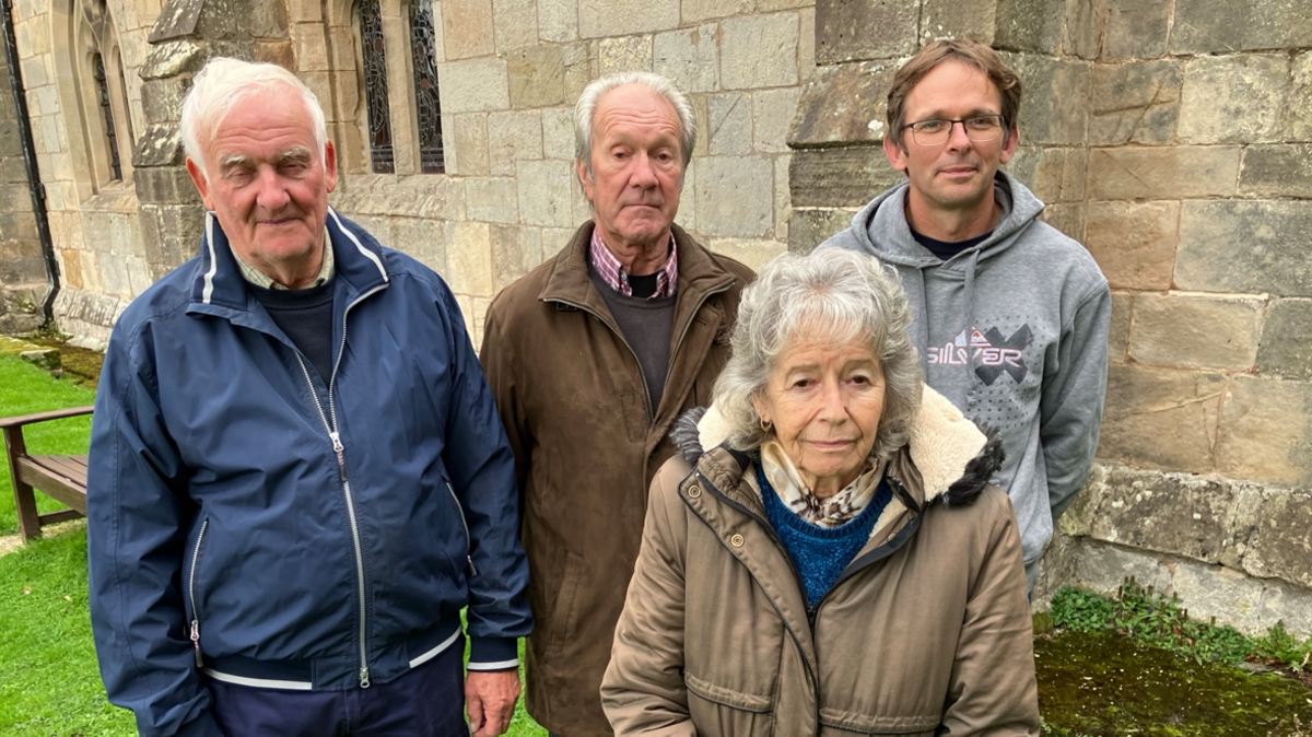 Three men and one woman standing on grass in front of the stone wall of a church, one man is wearing a blue jacket, one has a brown jacket and the other a pale grey hoodie. The woman is wearing a light brown coat.