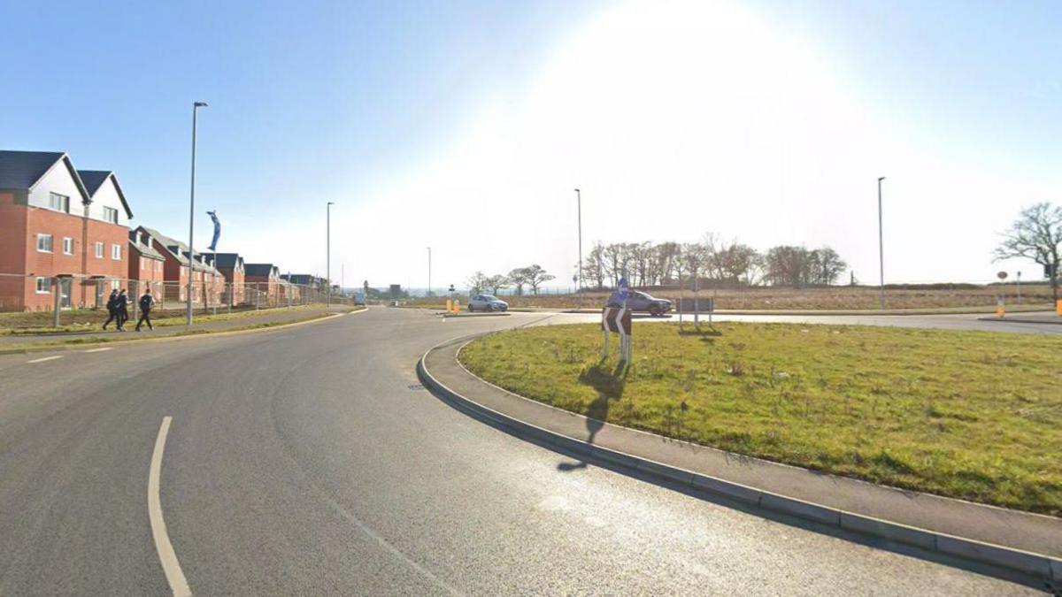 A roundabout, a tarmac road with some houses in the distance.