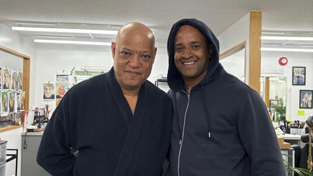 Laurence Fishburne stands inside room full of mirrors with pictures of people's faces on the wall and wearing a navy robe. He stands next to a mna