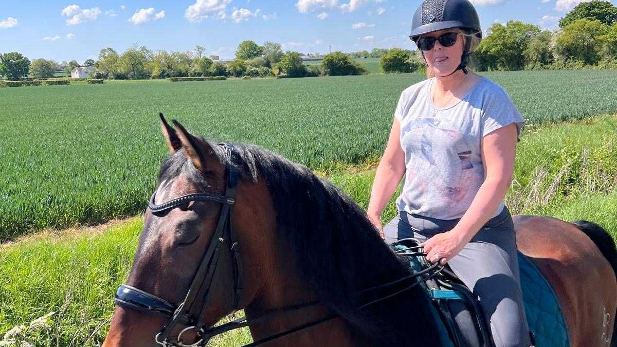 Mandy Young rides a bay coloured horse through a green field on a sunny day. She wears a black riding helmet, grey T-shirt, blue jodhpurs and sunglasses.