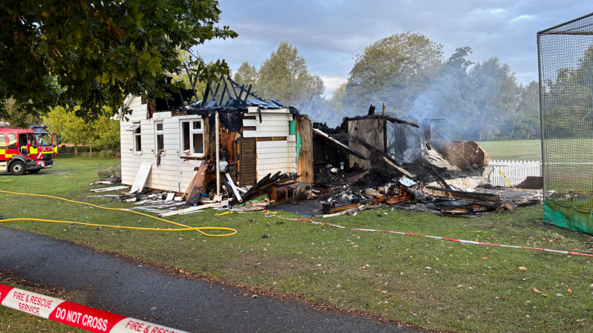 A white building is in the middle of the image on a field. A fire has made half of the building collapse, including its roof, which has left a rubble of wood on the right.