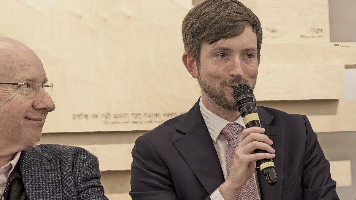 A seated Dan Tomlinson speaks into a microphone at the Finchley Reform Synagogue as Danny Finkelstein looks on