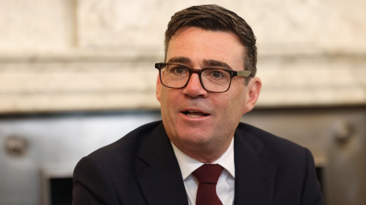 Andy Burnham, a man in a black jacket, with a white shirt and red tie. He has dark hair and is wearing black glasses.
