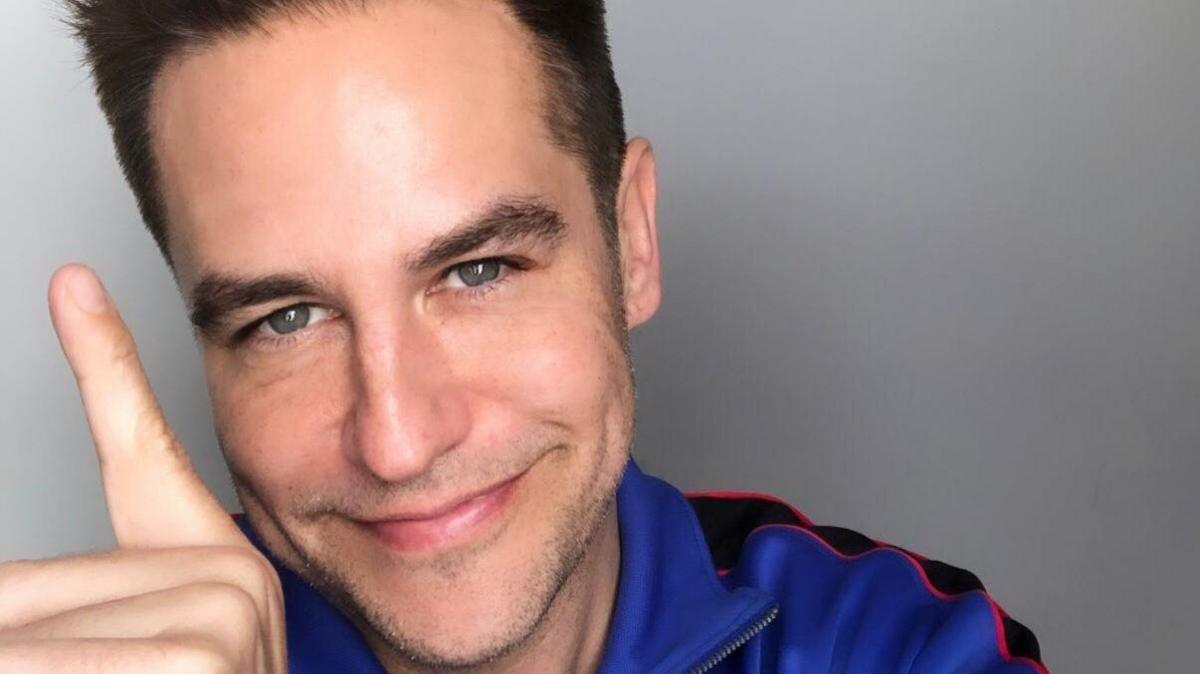 Anthony Kavanagh smiles as he holds an index finger up to celebrate one year of sobriety. He has dark hair and is wearing a blue top.