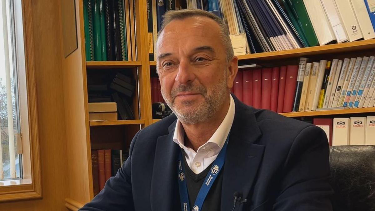 Guernsey Electricity chief executive Alan Bates sits on a leather chair in an office. A bookshelf filled with books and folders is behind him. Alan has short greying hair and a stubbly grey beard. He is wearing a dark blazer, dark jumper and white shirt. He has a blue lanyard round his neck with Guernsey Electricity's logo on it.