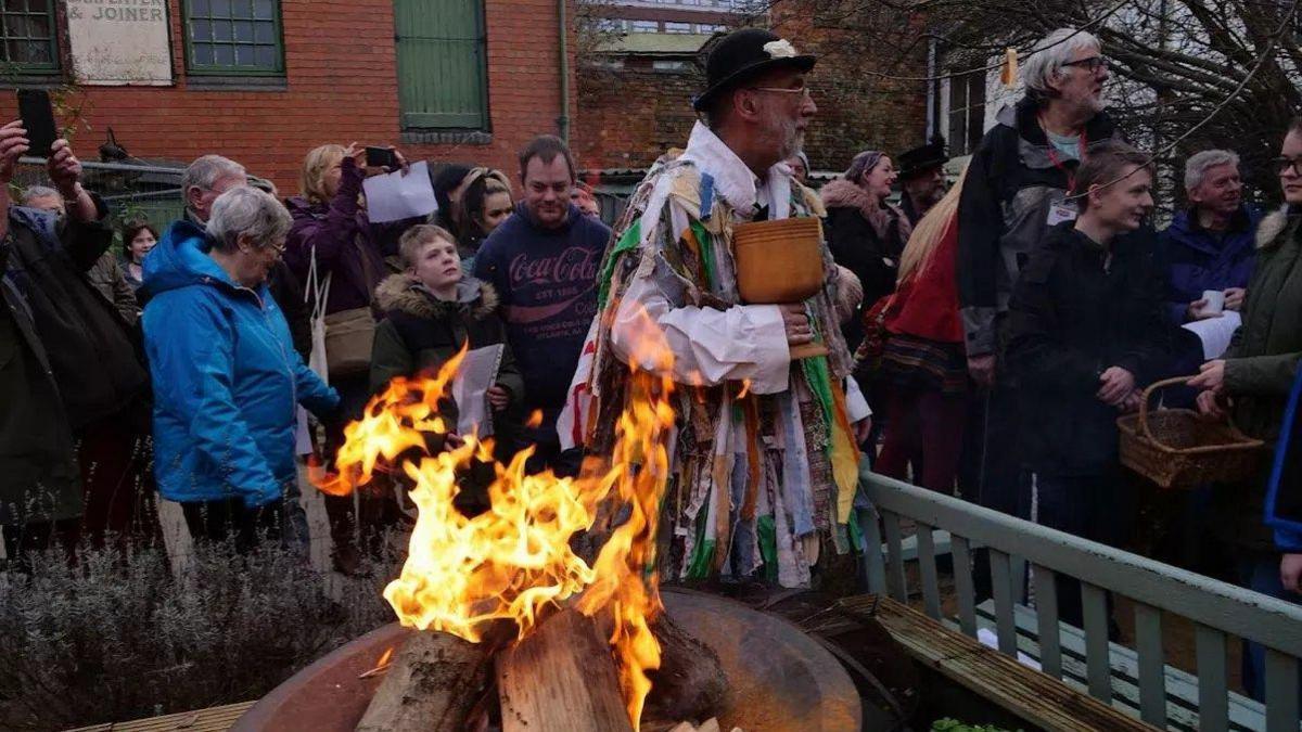 Bill Taylor dressed in a 'tatter' coat made from coloured strips of ribbons. He is surrounded by people as he leads a wassail event. He is stood next to a fire and is holding a large goblet.