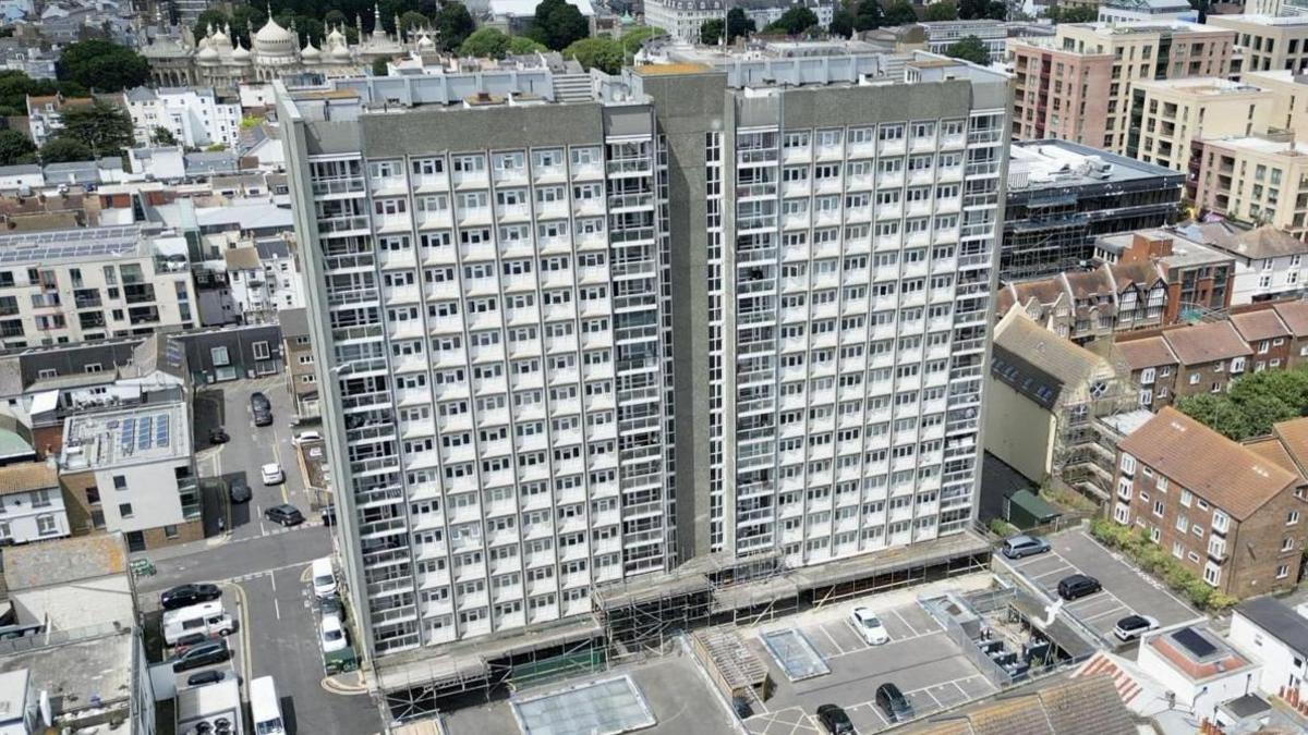 Two large grey tower blocks in an urban area, with a large but mostly empty car park at the front. Behind the building, in the distance, is the Brighton's Royal Pavilion