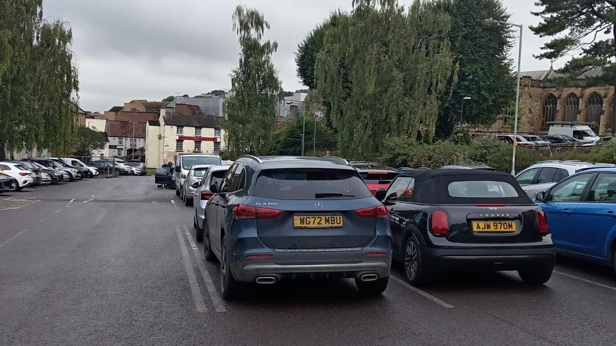 A car park in Yeovil. It is full of parked cars. A blue Mercedes and a black Mini Cooper are at the forefront of the picture. Buildings can be seen in the distance.