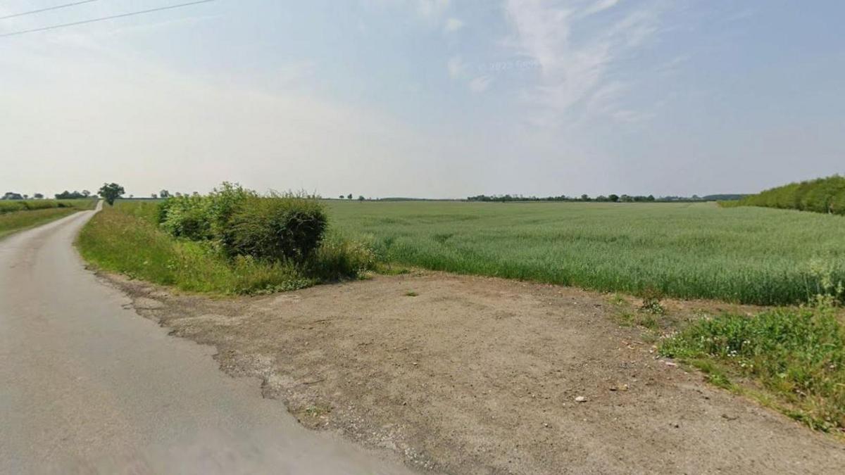 A Google street view of a country road with green fields to the right. The sky is blue with light clouds.
