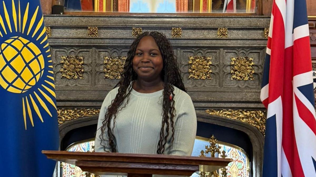 Zahara stands at the front of a room behind a small wooden desk. She is standing between two flags; the Union Jack and the Commonwealth of Nations flag which has a gold globe on a blue background.