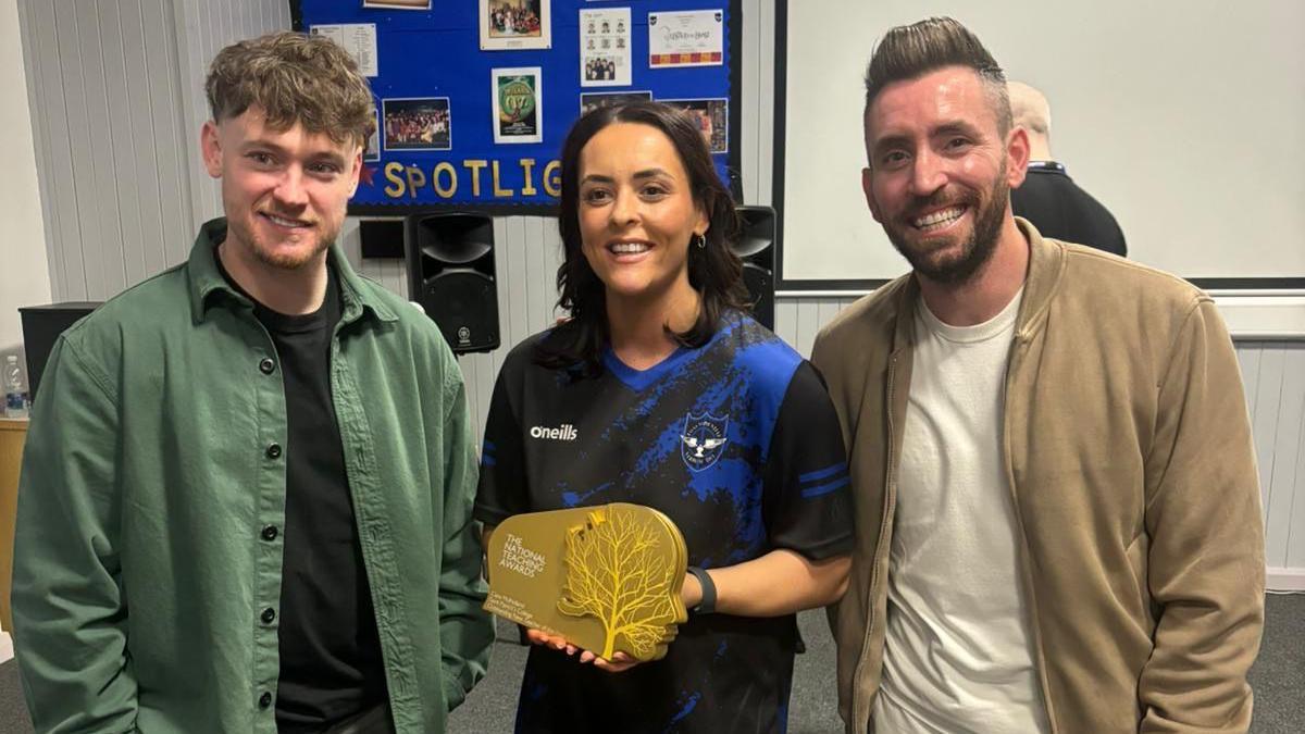 Ciara Mulholland, who has dark hair, and is wearing a school tracksuit, stands in between singer ryan mcmullan and One Show presenter Ryan Hand. She is holding an award.