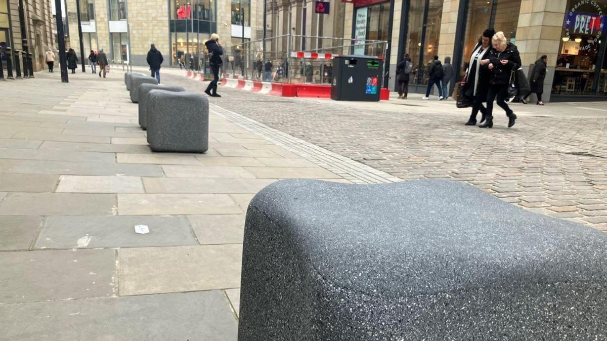 Stones in the shape of cubes on a street in Bradford are to be used as seats. Image shows road works on a shopping street and people walking around. 