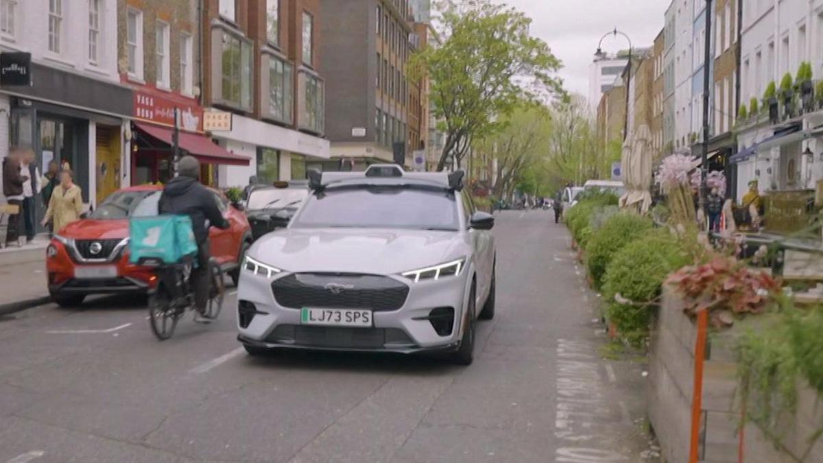 A white Ford Mach-E is shown driving down a street in London. A Deliveroo courier is cycling past the left side of car in the opposite direction. The right side of the road is flanked by bushes and flower boxes, which form a barrier between chairs and tables of restaurants' outdoor dining areas on pavements. The BT Tower is visible in the very background.