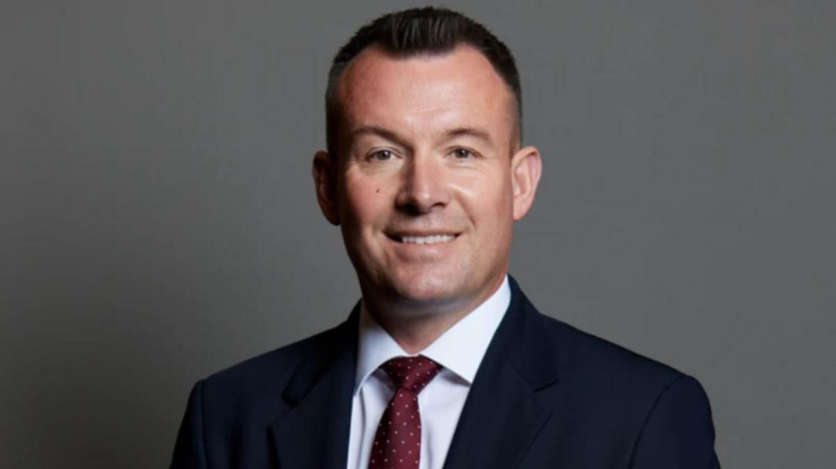 A man with short dark hair, wearing a dark suit, white shirt and red tie against a grey background