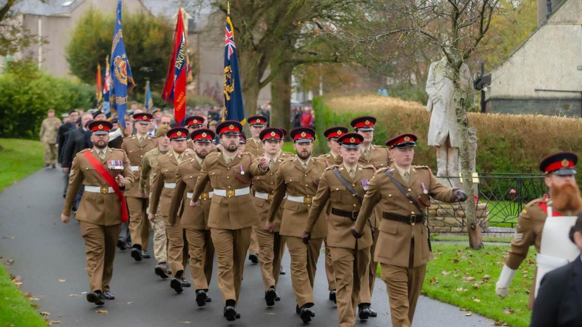 A parade, made up of men in a brown military uniform with black caps, marching along a path at Vulcan Park, Workington. Some are holding flags and the crowd can be seen following them in the blurred background.