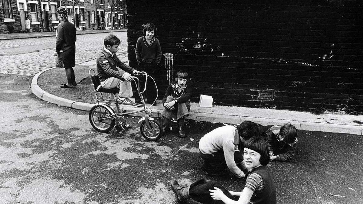 A black and white photo of six children playing on a street coroner while a woman with rollers in her hair stands on the pavement.