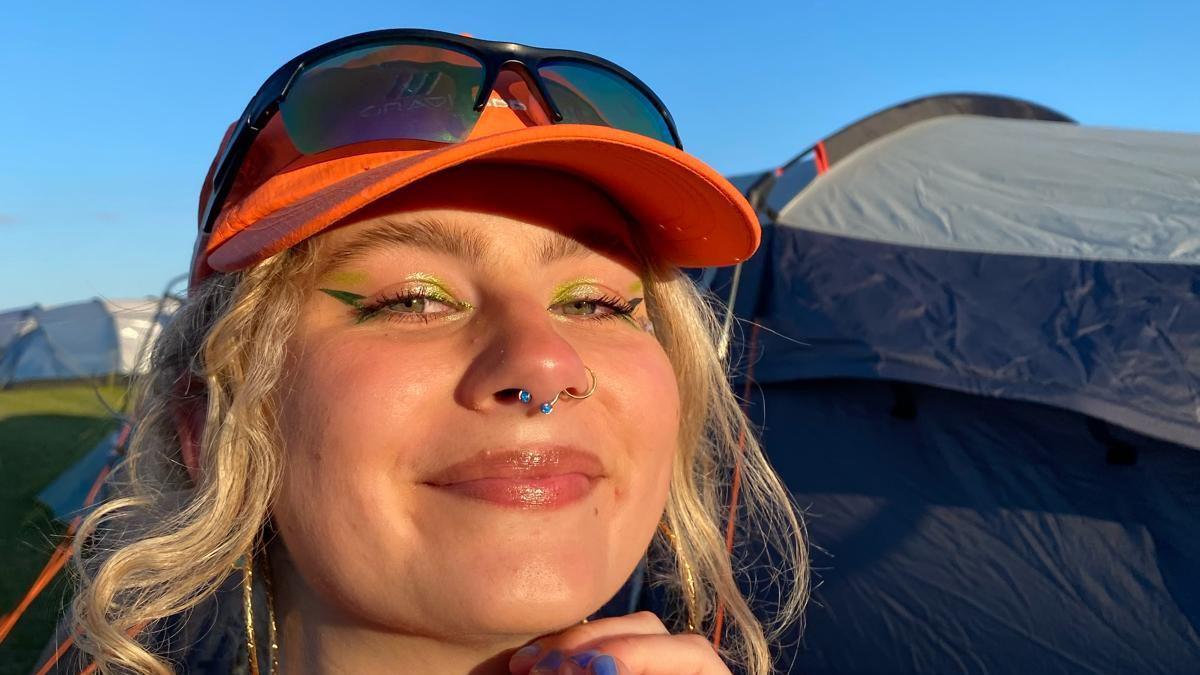 A woman wearing an orange hat and with colourful eye make-up smiles with the sun shining on her face