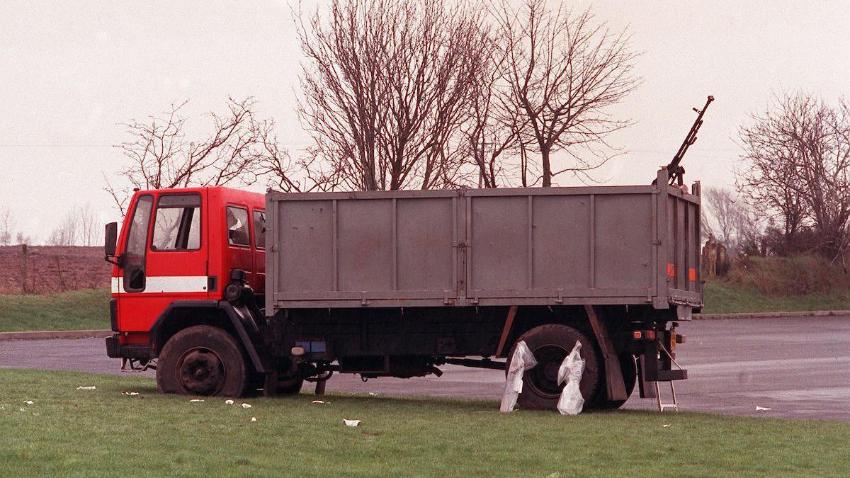 A red lorry with a grey carriage. Attached to the tailgate of the lorry is a heavy black machine gun, pointed to the sky. The lorry is sitting on a patch of grass beside a carpark.  