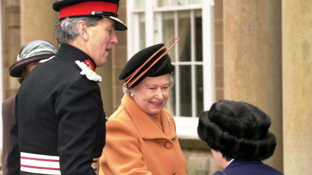 An archive shot of Elizabeth II in 2000, she is wearing an orange coat and a black and orange hat. To her right is a tall man in a dark, formal military uniform, to her left is a woman, seen from behind, wearing a dark blue jacket and fur hat.