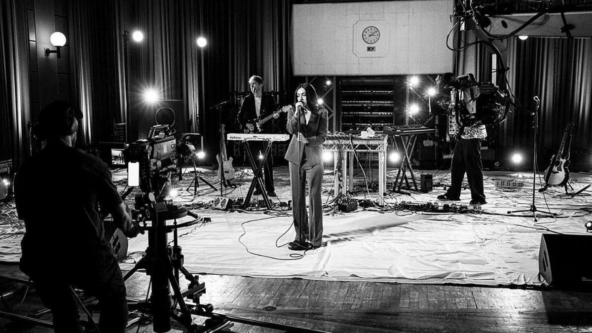 A black and white photo of Charli XCX performing at BBC Maida Vale studios. She is wearing a suit, and there are two people performing behind her. On the left hand side of the photograph, you can see the back of a camera person and a camera filming the performance. 