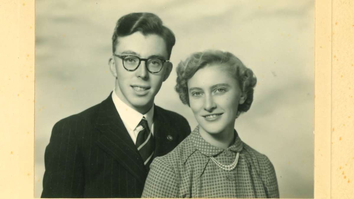 Mary, on the right, wearing checked blouse with what looks like a double string pearl necklace. Her hair is short and styled. She is with her husband Philip, on the left, wearing round glasses and a suit jacket, shirt and tie. They are both looking at the camera, in a posed photograph. The image is in black and white.
